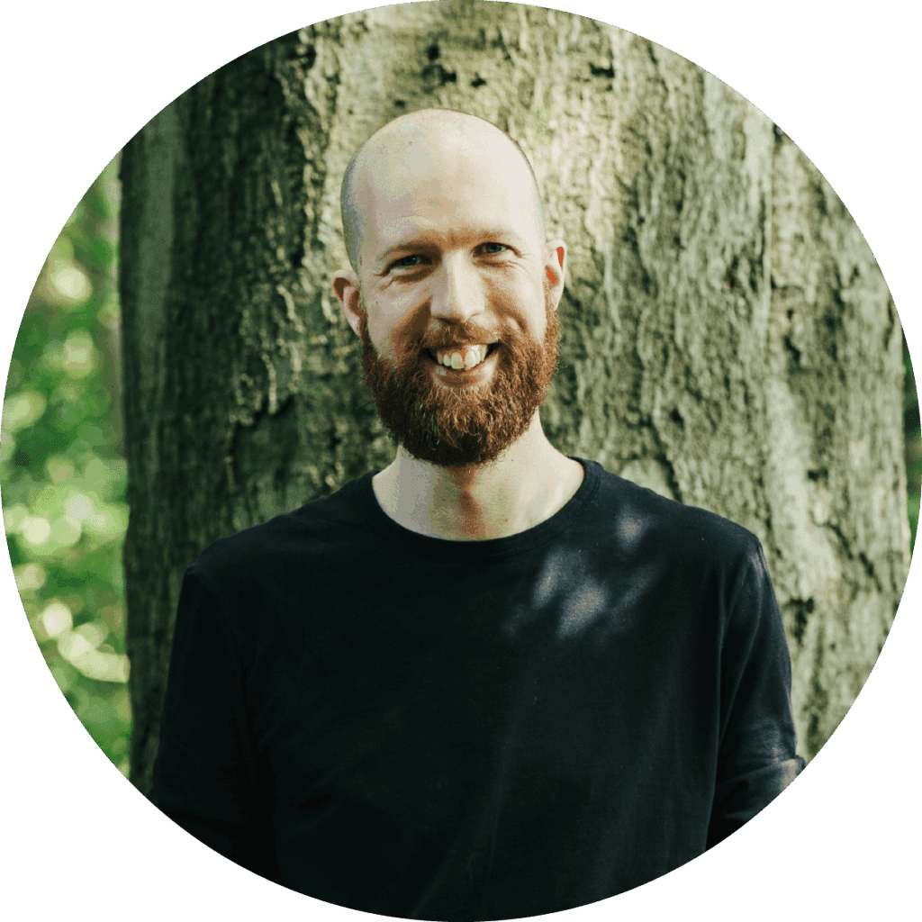Picture of Thom Baker smiling infront of large tree or forest background